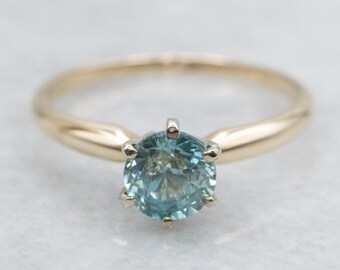 May include: A gold ring featuring a round, light blue gemstone. The gemstone is set with six prongs, and the band is a simple, polished gold. The ring is displayed on a white surface.