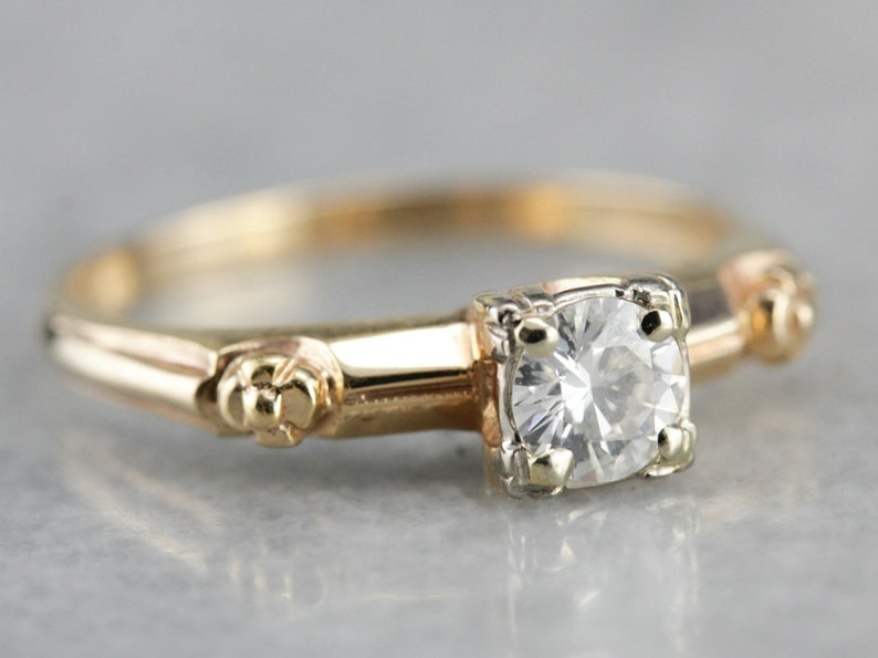 May include: A vintage engagement ring with a single round diamond set in a gold band. The band is decorated with small floral details.