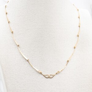 May include: A delicate gold necklace with a minimalist design. The necklace features slender, curved bars connected by small, round beads. The clasp is a simple, heart-shaped design. The necklace is displayed on a white surface.