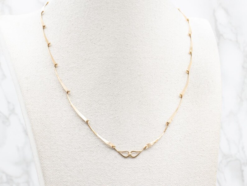 May include: A delicate gold necklace with a minimalist design. The necklace features slender, curved bars connected by small, round beads. The clasp is a simple, heart-shaped design. The necklace is displayed on a white surface.