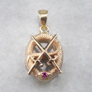 Puede incluir: Colgante dorado con un diseño detallado de una serpiente que rodea dos espadas cruzadas. Una pequeña gema roja redonda está engastada en la base de la serpiente. El colgante tiene una superficie texturizada y un lazo para una cadena.