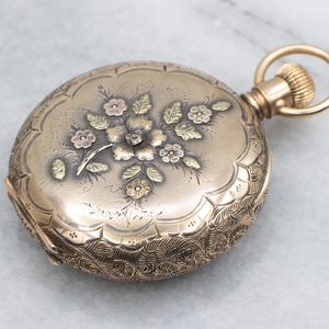 May include: A gold pocket watch with an ornate floral design. The watch face features a detailed engraving of flowers and leaves in shades of gold and pink. The watch has a winding stem and a circular bail.