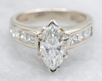 Puede incluir: Anillo de oro blanco con un prominente diamante talla marquesa. La banda está engastada con diamantes más pequeños de talla cuadrada a cada lado de la piedra central. El anillo se muestra sobre una superficie blanca.