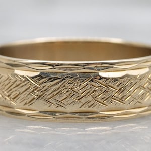 May include: A gold band ring featuring a central textured pattern of diagonal, crisscrossing etched lines. The ring is bordered on the top and bottom edges by a series of repeating, elongated diamond-shaped facets that catch the light.