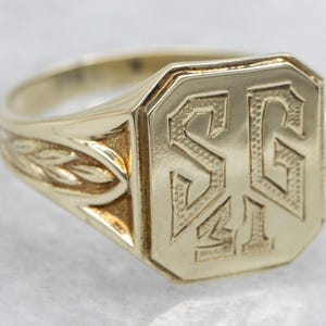 May include: A gold signet ring with an octagonal face. The face is engraved with the initials "SG" and "I". The band features a leaf design on the side.