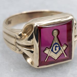 May include: A gold-toned ring with a rectangular, burgundy-colored center stone. The stone features a Masonic symbol with a compass, square, and the letter "G" in blue and gold. The ring has a ribbed band.