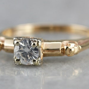 May include: A gold ring with a single round diamond set in a prong setting. The ring has a simple band with small decorative accents on either side of the diamond.