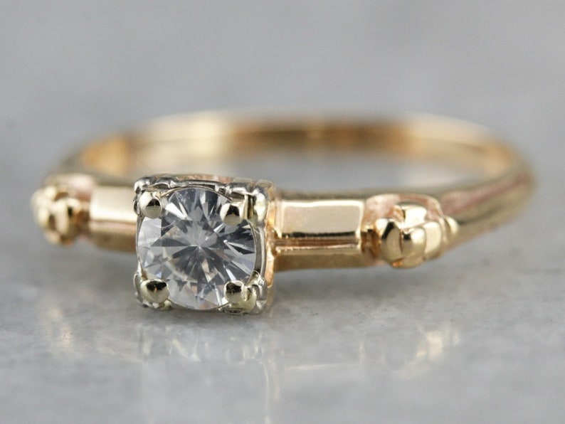 May include: A gold ring with a single round diamond set in a prong setting. The ring has a simple band with small decorative accents on either side of the diamond.