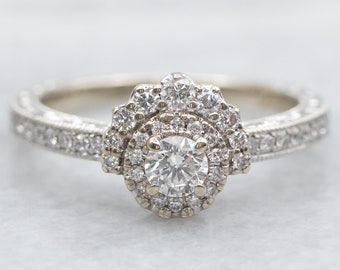 May include: A delicate diamond engagement ring with a round center stone and a double halo of smaller diamonds. The band is also set with smaller diamonds in a milgrain design.