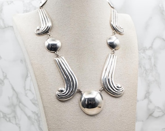 May include: A silver necklace with a large, round pendant and smaller, round accents. The necklace is made of a series of curved, textured pieces.