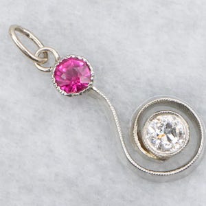 Op de afbeelding: Een zilveren hanger met een helder roze edelsteen en een diamant in een wervelend ontwerp. De hanger heeft een lus voor een ketting. De edelstenen zijn rond en gezet in een bezel.