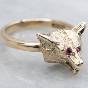 May include: Gold-toned ring featuring a wolf head design. The wolf's head has textured details and is adorned with two small, round, ruby-colored gemstones for eyes. The ring is a jewelry item.