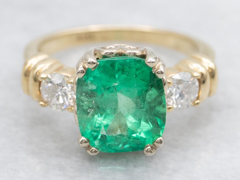 May include: A gold ring featuring a large, vibrant green emerald center stone, flanked by two smaller, sparkling white diamonds. The ring has a classic design with a polished gold band and prong settings.