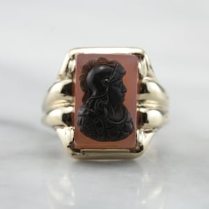 Puede incluir: Un anillo de oro con una piedra de cornalina rectangular con un camafeo negro de una persona con casco.