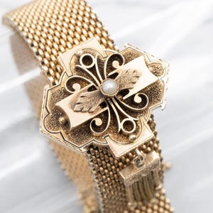 May include: A gold mesh bracelet with a decorative clasp featuring a cross design with a pearl accent.