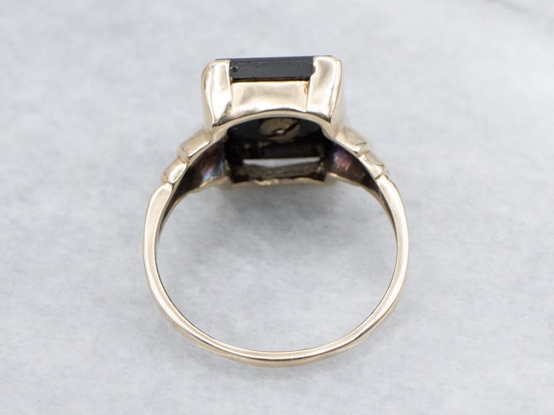 May include: A gold ring featuring a large, rectangular black gemstone. The ring has a classic design with decorative elements on the shoulders and a smooth, polished band. The gemstone is set in a secure bezel setting.