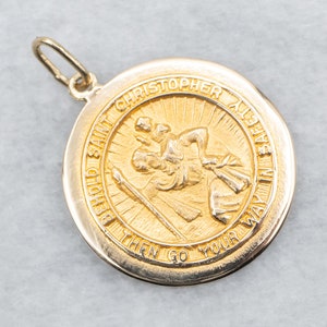 Op de afbeelding: Gouden hanger met een afbeelding van Sint Christoffel die een kind draagt. De hanger heeft de inscriptie "Behold Saint Christopher All Safe In Thy Way Then Go Your Way In Safety".