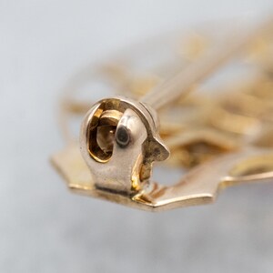 May include: Close-up of the back of a gold-toned brooch with a pin and catch closure.