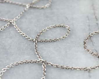 May include: Close-up of a silver-colored chain necklace with small, oval links. The chain is arranged in a loose, flowing pattern against a light gray background. The links are uniform in size and shape, creating a delicate and elegant appearance.