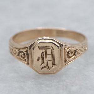 May include: A gold-toned signet ring with an octagonal face. The face features an engraved letter "D" in a decorative font. The ring's shoulders are adorned with scrollwork. A classic piece of jewelry.