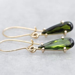 Peut inclure: Une paire de boucles d'oreilles pendantes dorées avec des pierres précieuses vertes foncées en forme de poire. Les pierres précieuses sont facettées et légèrement translucides. Les boucles d'oreilles ont un design simple en forme de crochet.