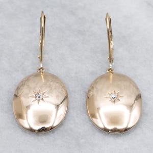 May include: A pair of gold-coloured oval drop earrings. Each earring has a central starburst design with a small, clear gemstone. The earrings have a lever-back closure and a polished finish.