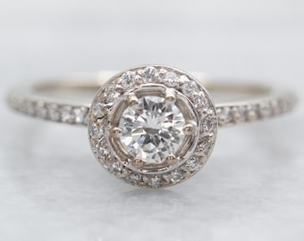 May include: A diamond engagement ring with a round center stone and a halo of smaller diamonds. The band is also encrusted with small diamonds. The ring is set in a white metal, likely platinum or white gold.