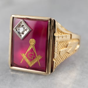 May include: A gold Masonic ring with a red stone featuring a gold Masonic symbol and a small diamond. The ring band is textured with a design of a ruler and a compass.