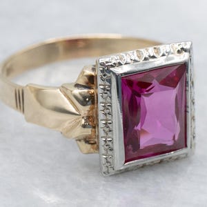 May include: A gold ring with a large, rectangular, magenta-colored gemstone. The gemstone is set in a silver-colored bezel with decorative detailing. The ring's band is gold and features a textured design near the gemstone setting.