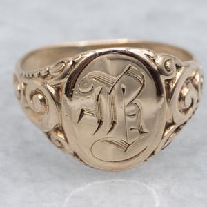 Op de afbeelding: Een gouden zegelring met een ovale voorkant, met een gegraveerde letter "R" in een sierlijk lettertype. De ring heeft decoratief krulwerk op de schouders en een gepolijste afwerking.