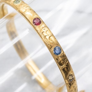 以下が含まれることがあります： 金色のバングルブレスレット。彫刻された花の模様とカラフルな宝石が特徴です。赤、青、緑の色合いの丸い宝石が並んでいます。このバングルは、クラシックなジュエリーです。