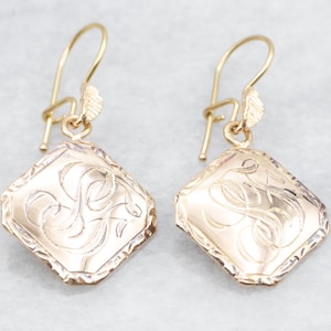 Puede incluir: Un par de pendientes de color dorado con un colgante cuadrado que presenta un monograma grabado. Los pendientes tienen un diseño simple y elegante.