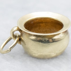 Peut inclure: Un petit charm doré poli en forme de chaudron ou de pot miniature. L'objet présente un corps arrondi avec un bord large et évasé, ainsi qu'un anneau de jonction circulaire fixé sur le côté, permettant de le suspendre comme un pendentif.