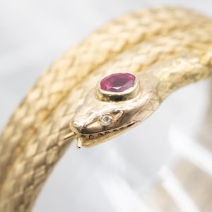 Op de afbeelding: Een gouden slangenarmband met een robijn edelsteen in de kop. De slangenkop heeft een kleine diamant accent en gedetailleerde schubben. De armband is een uniek sieraad.
