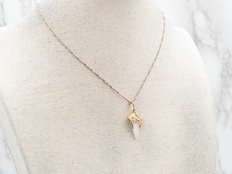 May include: A gold chain necklace with a white crystal pendant. The pendant is shaped like a pointed tooth with gold detailing.