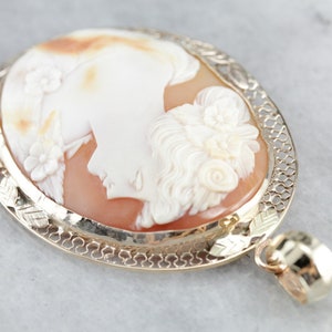 May include: A cameo pendant featuring a carved shell with a detailed profile of a woman's face. The cameo is set in a gold filigree frame with a delicate chain.