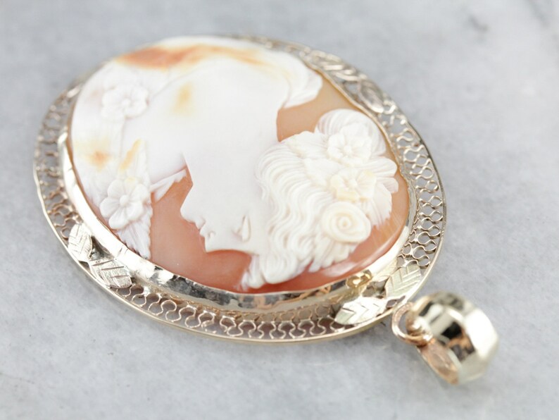 May include: A cameo pendant featuring a carved shell with a detailed profile of a woman's face. The cameo is set in a gold filigree frame with a delicate chain.