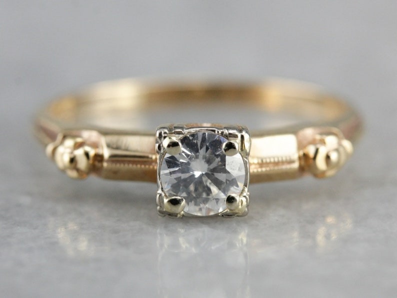 May include: A vintage gold engagement ring with a single round diamond set in a prong setting. The band features a delicate floral design.