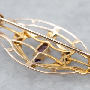 May include: A gold-toned oval-shaped brooch with a delicate filigree design and a purple gemstone in the center. The brooch has a pin and catch closure on the back.