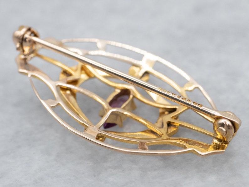 May include: A gold-toned oval-shaped brooch with a delicate filigree design and a purple gemstone in the center. The brooch has a pin and catch closure on the back.