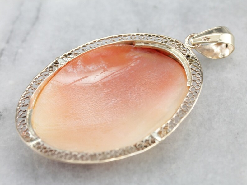 May include: A delicate silver filigree pendant featuring a large oval shell cameo. The cameo is a pale pink color with a smooth, polished surface.