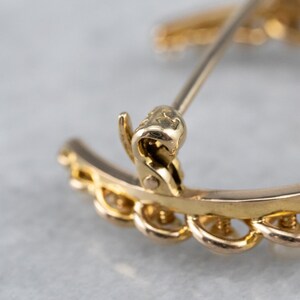 Puede incluir: Un broche de tono dorado en forma de media luna con un dise&ntilde;o detallado. El broche tiene un cierre de pin y enganche y est&aacute; marcado con la letra "K".