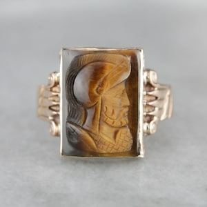 May include: A gold ring with a carved tiger's eye stone featuring a profile of a person wearing a helmet.