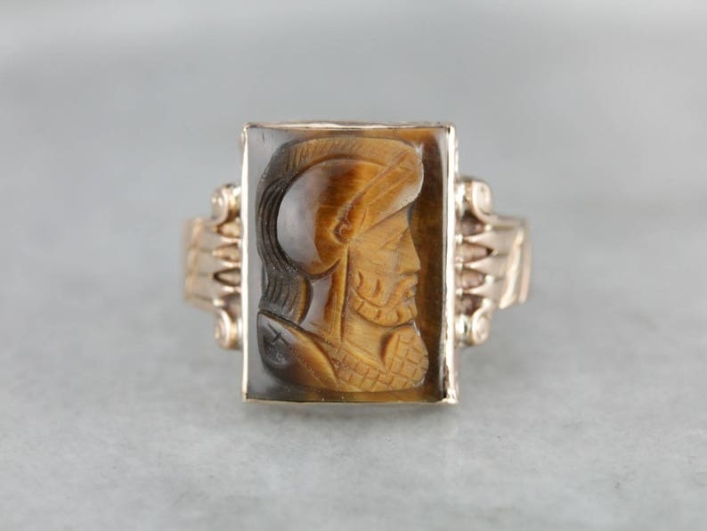 May include: A gold ring with a carved tiger's eye stone featuring a profile of a person wearing a helmet.