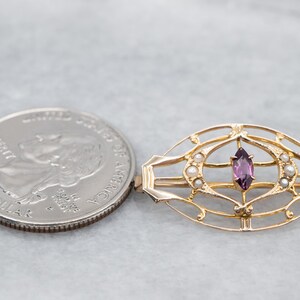 May include: A gold-toned brooch with a delicate, intricate design featuring a purple gemstone surrounded by small white pearls.