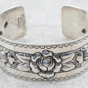 Puede incluir: Pulsera de brazalete de plata con un diseño floral con rosas y hojas. La pulsera está grabada con un borde festoneado.