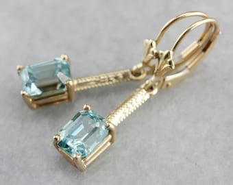 May include: A pair of gold earrings with light blue emerald-cut gemstones. The earrings have a delicate, ornate design and dangle from a leverback closure.
