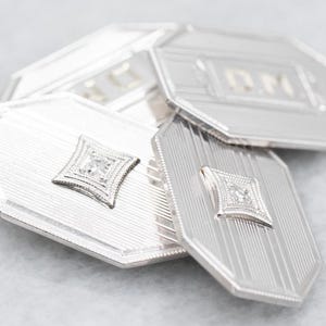 Könnte beinhalten: Nahaufnahme eines Sets silberner Manschettenknöpfe. Jeder Manschettenknopf hat eine geometrische Form mit einem rautenförmigen Mittelstein. Die Manschettenknöpfe haben ein strukturiertes, gestreiftes Design.