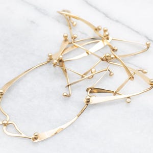 May include: A gold-toned necklace with an abstract design. The necklace features interconnected, curved metal pieces with small, round accents. The overall shape is irregular and organic, suggesting a modern or artistic style. The metal has a polished finish.
