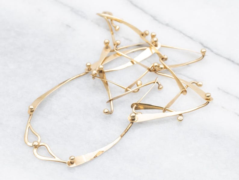 May include: A gold-toned necklace with an abstract design. The necklace features interconnected, curved metal pieces with small, round accents. The overall shape is irregular and organic, suggesting a modern or artistic style. The metal has a polished finish.